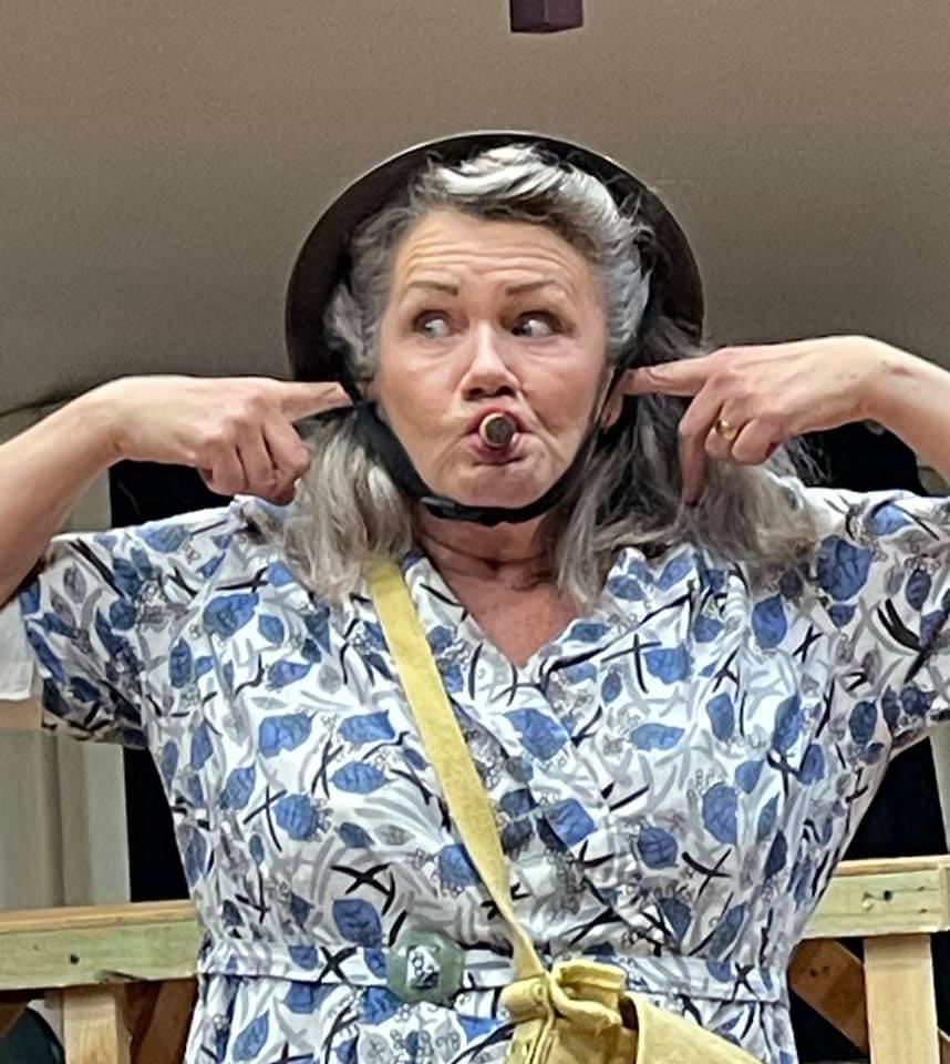 A middle aged woman in nineteen forties clothing wearing a army helmet. She has her fingers in her ears and a cork in her mouth.